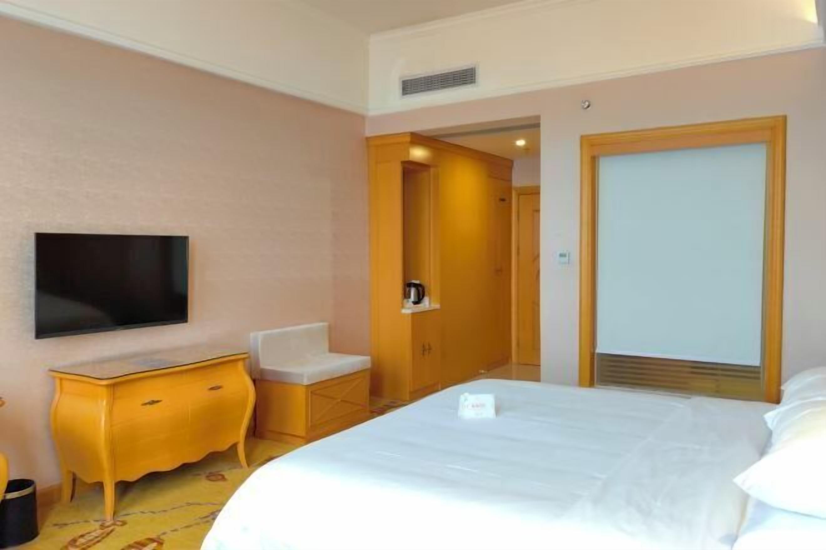A hotel room with a bed, a TV, a wooden wardrobe, a small desk, and a chair.