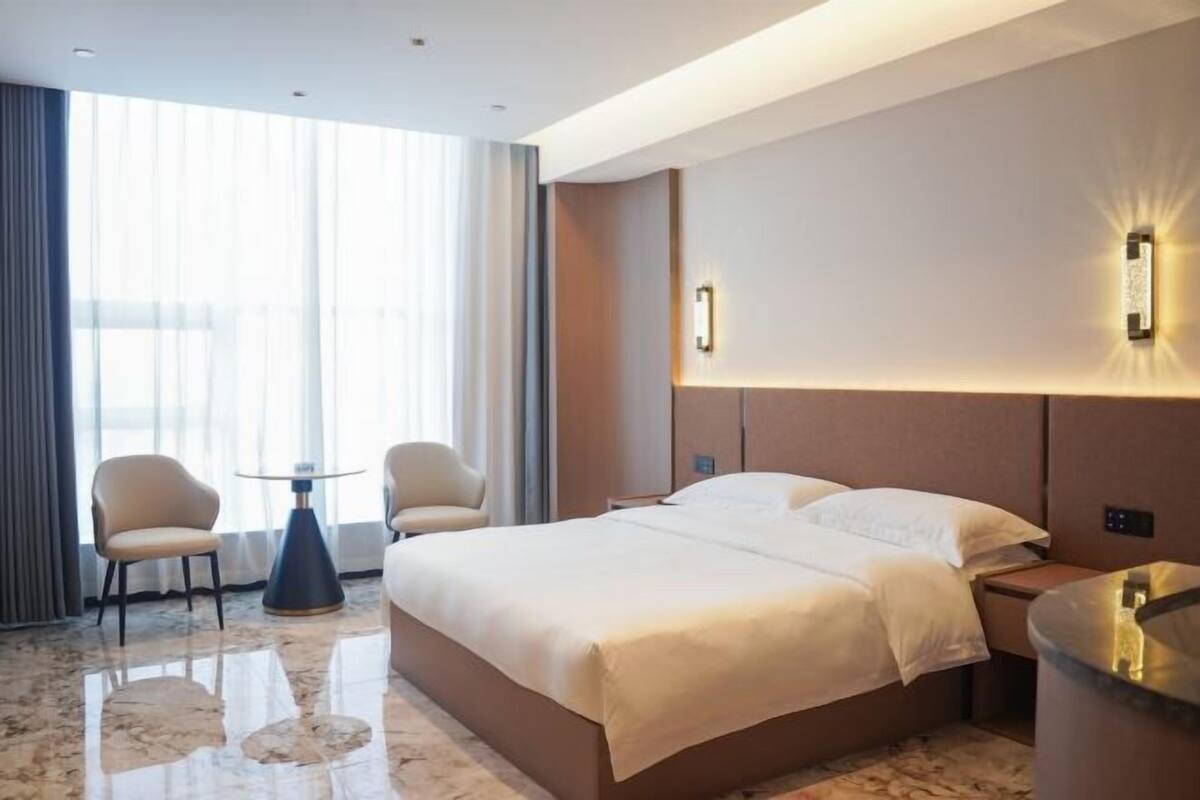 A hotel room with a bed, two chairs, a small table, and a wall-mounted light.