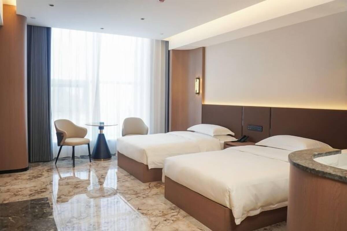 A hotel room with two beds, a small table, a chair, and a large window.
