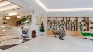 A modern reception area with a clean, white floor, a reception desk, and a bookshelf filled with books and plants. There are two green chairs and a small table in the center. - GreenTree Inn Smart Choice Hotel (Changzhou Xixiashu Branch) (Huludao)