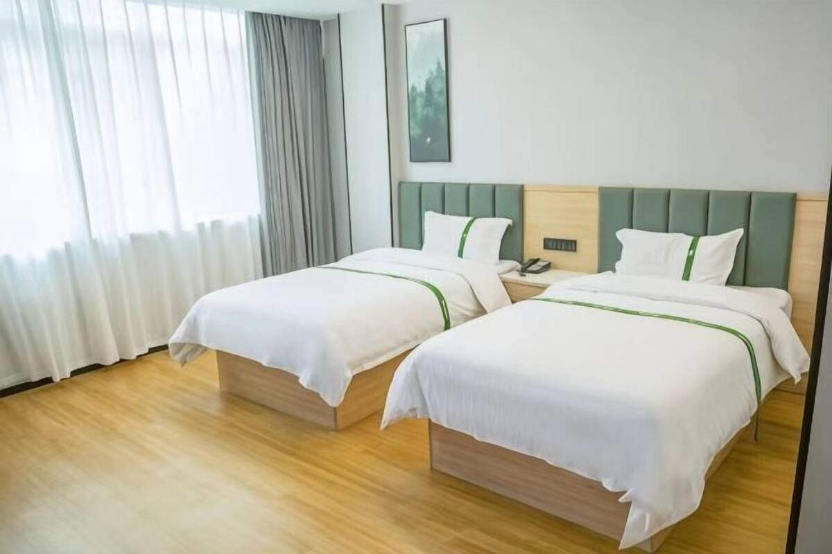 A hotel room with two beds, wooden flooring, a nightstand, and a window with curtains.