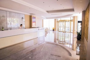A hotel reception area with a reception desk, a waiting area with chairs, and a large window providing natural light.