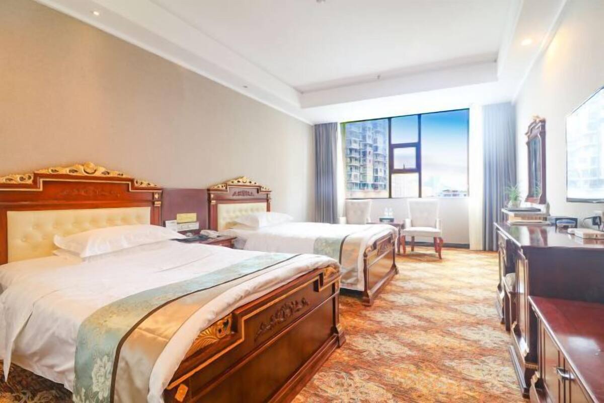 Ein Hotelzimmer mit zwei Betten, einem Schreibtisch, einem Stuhl und einem großen Fenster mit Blick auf die Stadt.