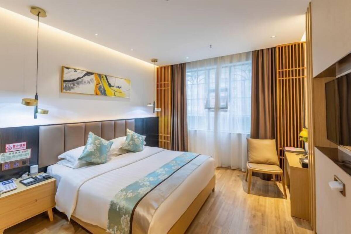 Ein Hotelzimmer mit Bett, Nachttisch, Sessel, Schreibtisch, Fernseher, Kunstwerk und Vorhängen.