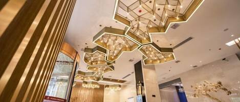 A modern hotel lobby with a geometric ceiling design, a reception desk, and a large window.