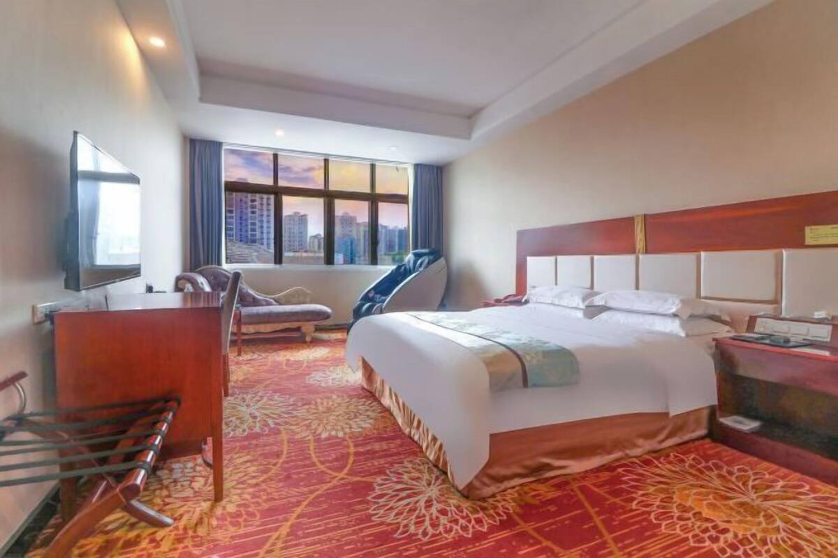 Ein Hotelzimmer mit einem großen Bett, einem Schreibtisch, einem Sessel, einem Fernseher und einem Fenster mit Blick auf die Stadt.