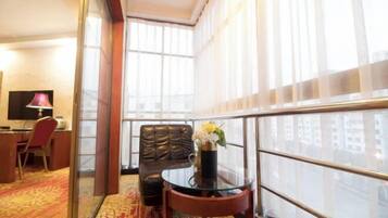 A hotel room with a large window, a leather chair, a glass table with a vase of flowers, and a desk with a lamp.