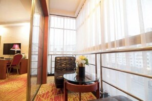 Une chambre d’hôtel dotée d’une grande fenêtre, d’un fauteuil en cuir, d’une table en verre ornée d’un vase de fleurs et d’un bureau avec une lampe.