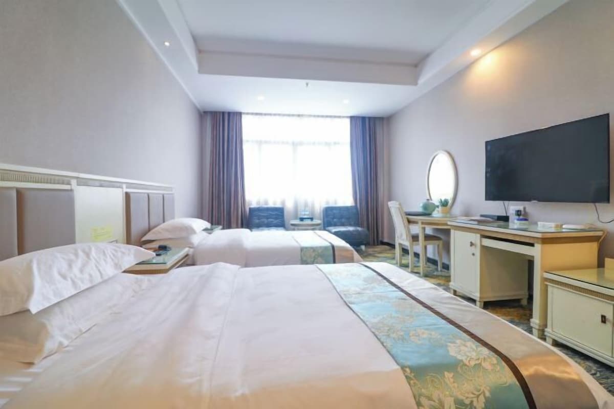 Une chambre d’hôtel avec deux lits, un bureau, une chaise, un téléviseur à écran plat et une grande fenêtre avec des rideaux.