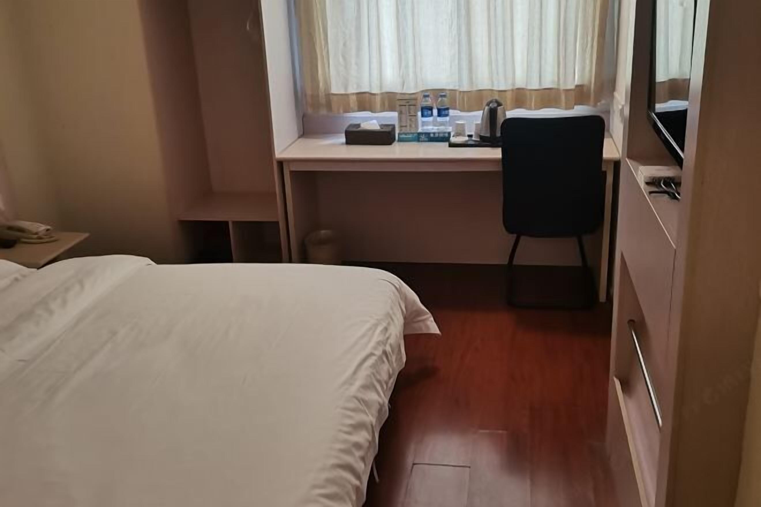 A hotel room with a bed, a desk with a chair, a window with curtains, and a bedside table with a lamp.