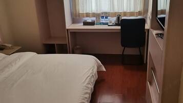 A hotel room with a bed, a desk with a chair, a window with curtains, and a bedside table with a lamp.