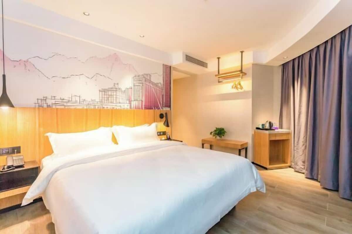 A hotel room with a large bed, a nightstand, a wall-mounted lamp, a small table with a plant, and a window with curtains.