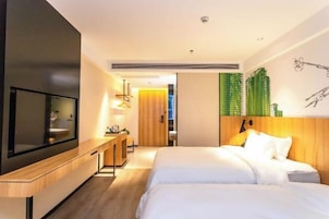A modern hotel room with a large flat-screen TV, a wooden desk, a bed with white bedding, and a green wall mural.