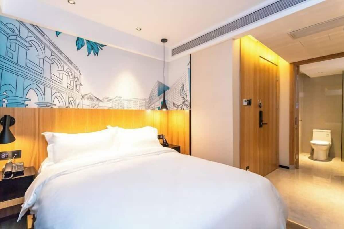 A hotel room with a large bed, a wooden headboard, a nightstand with a lamp, a wall mural, and a wooden door leading to a bathroom.
