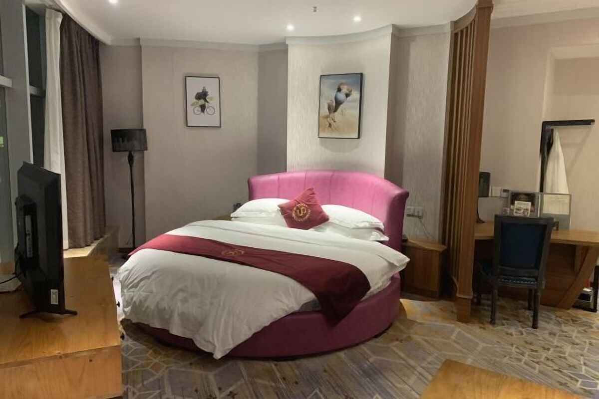 A circular bed with a pink headboard and white bedding, a desk with a chair, a lamp, and artwork on the walls.