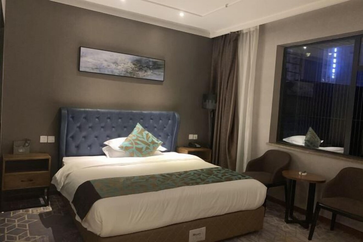 A hotel room with a large bed, a nightstand, a chair, a small table, and a window with curtains.