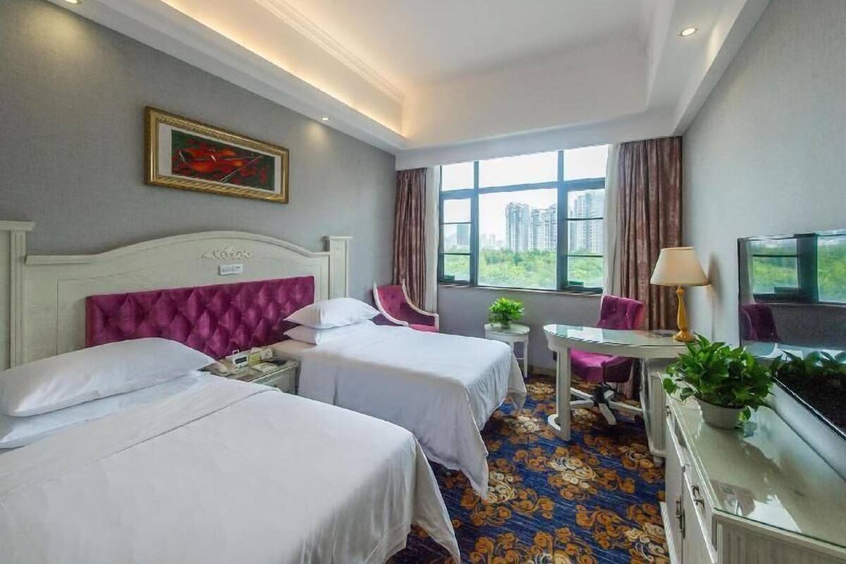 A hotel room with two beds, a desk, a chair, a TV, and a window with a city view.