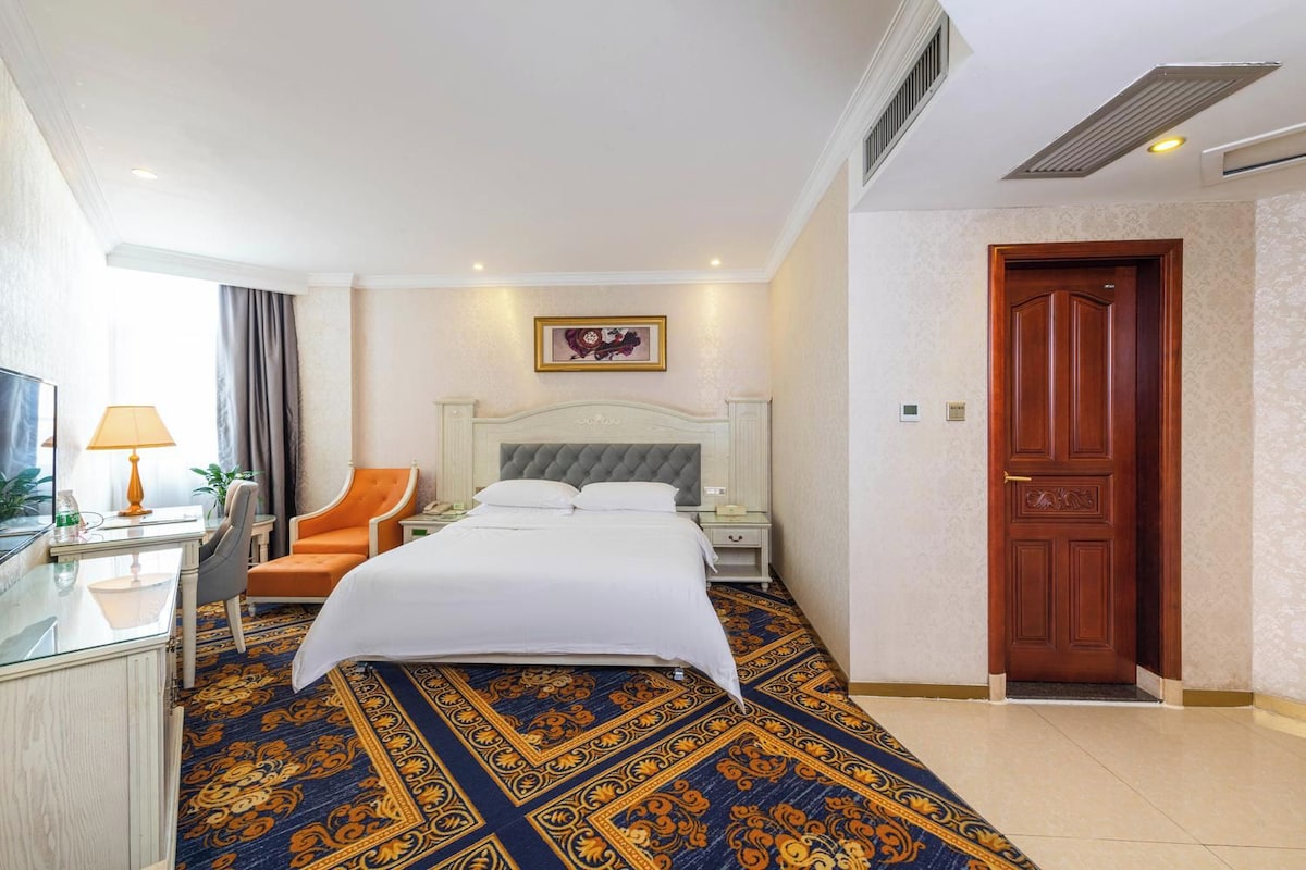 A hotel room with a bed, a desk with a lamp, an orange chair, a wooden door, and a patterned carpet.