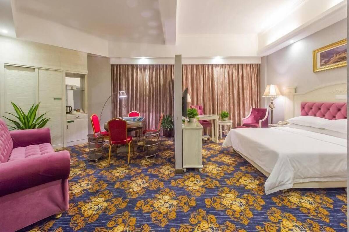 A hotel room with a bed, a desk with a chair, a small table with a chair, a potted plant, and a large window with curtains.