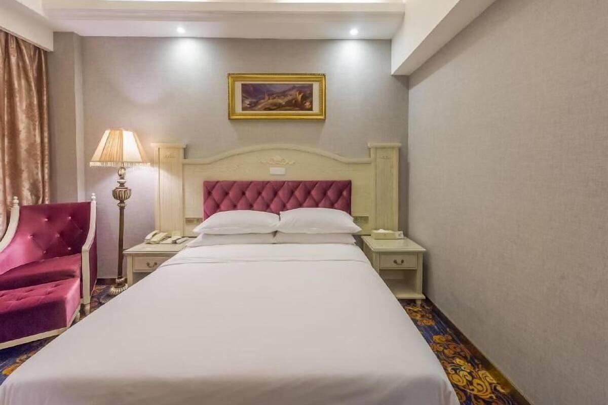 A hotel room with a large bed, a red armchair, a nightstand with a lamp, and a painting on the wall.