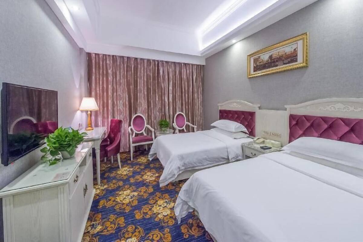 A hotel room with two beds, a desk with a lamp, a chair, a television, and a patterned carpet.
