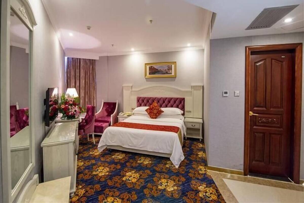 A hotel room with a large bed, a sitting area with a chair, a desk with a lamp, and a door.