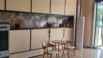 Een kamer met een groot raam, een wandairconditioner, een klein tafeltje met stoelen en een plank met potplanten.