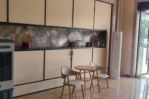 Una habitación con un ventanal grande, un aire acondicionado de pared, una mesa pequeña con sillas y una estantería con plantas en macetas.