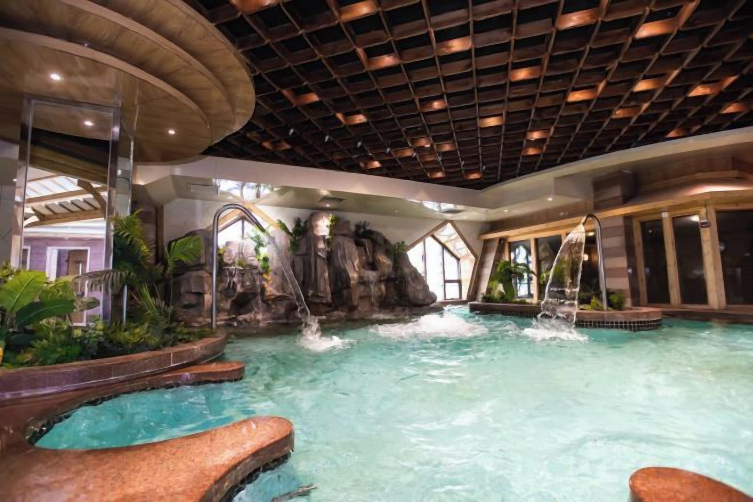 An indoor swimming pool with a waterfall feature, surrounded by tropical plants and a modern ceiling design.