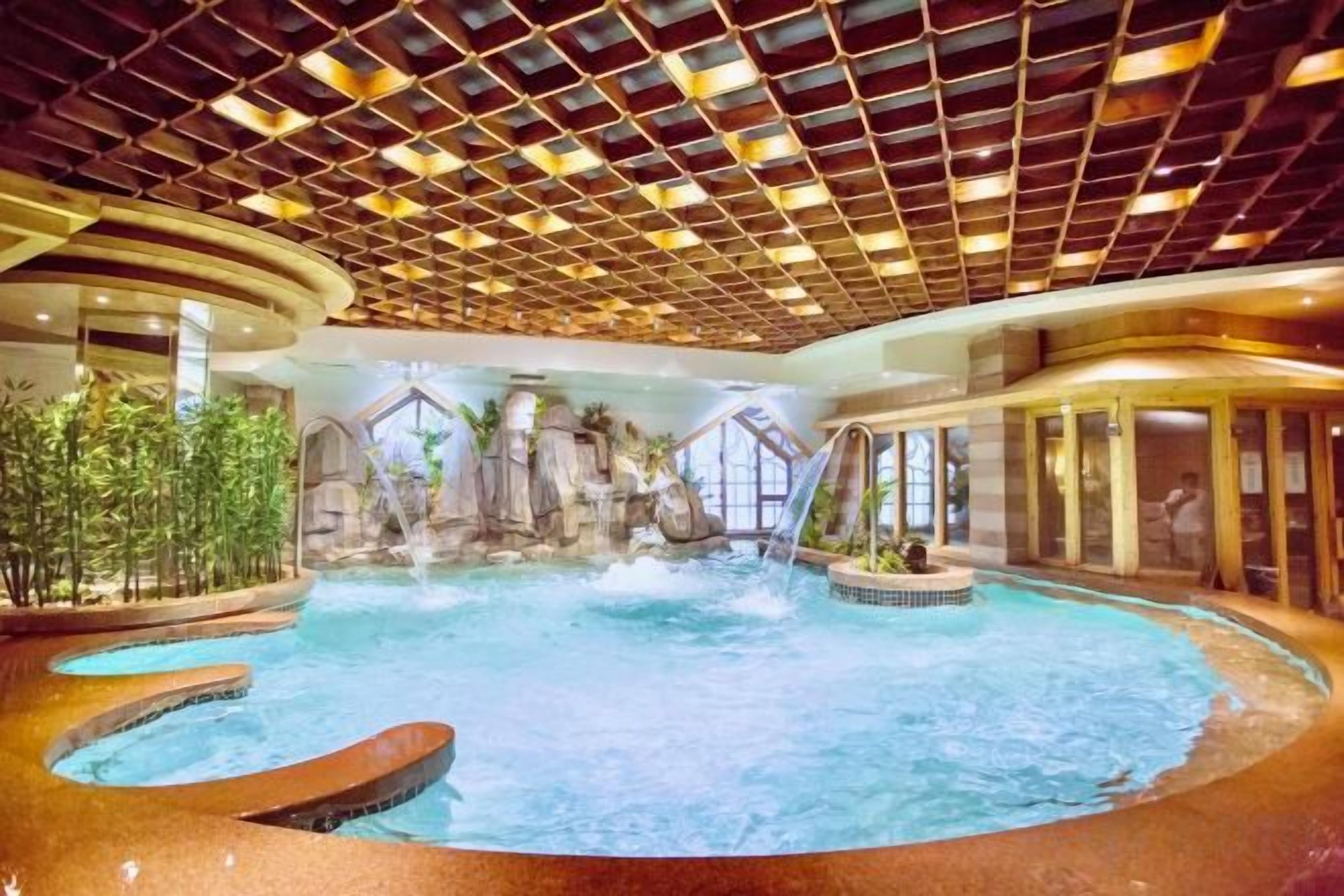 Una amplia piscina interior con caída de agua, rodeada por un techo alto con diseño de cuadrícula y un fondo de pared de piedra.