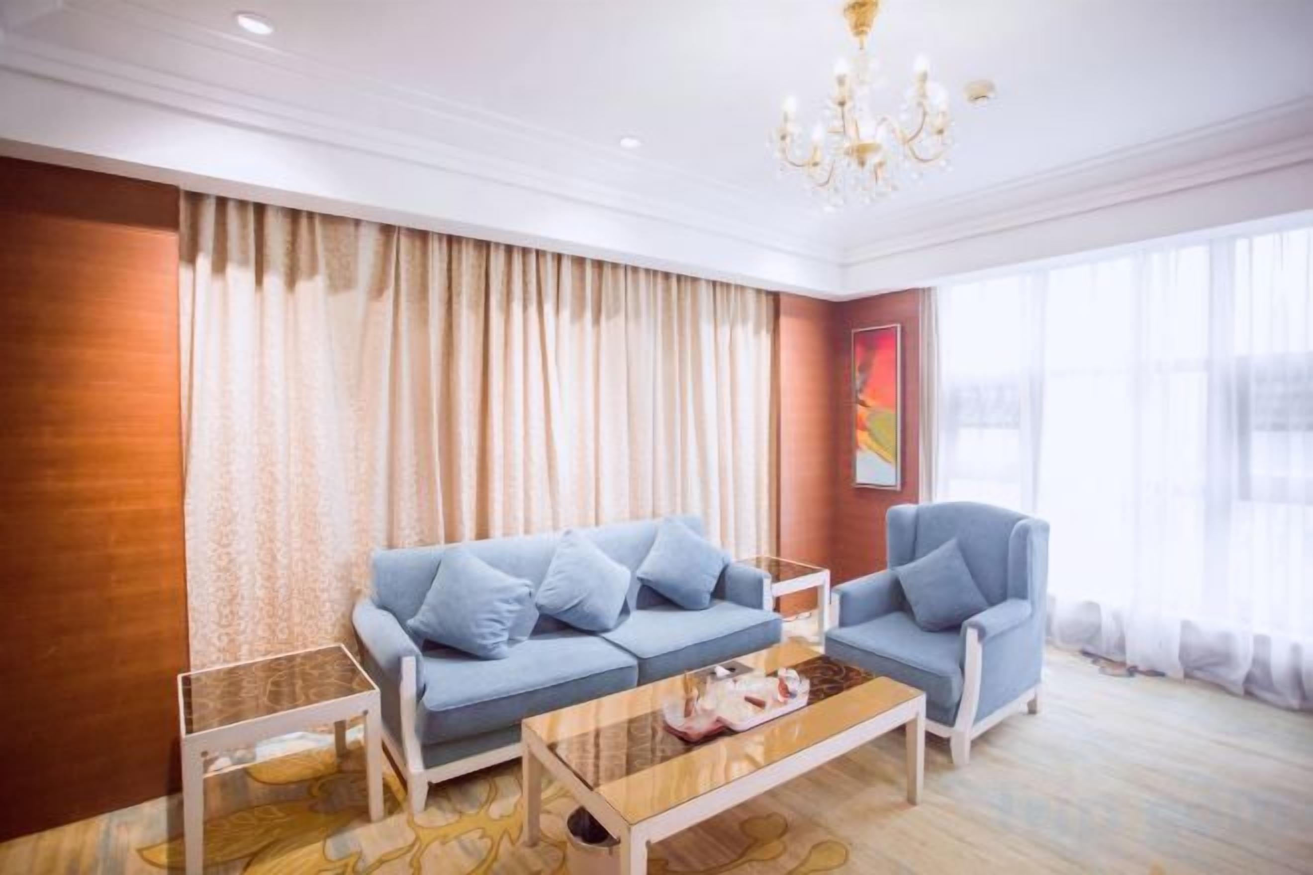 A modern living room with a sofa, armchair, and coffee table, a chandelier, and large windows with curtains.