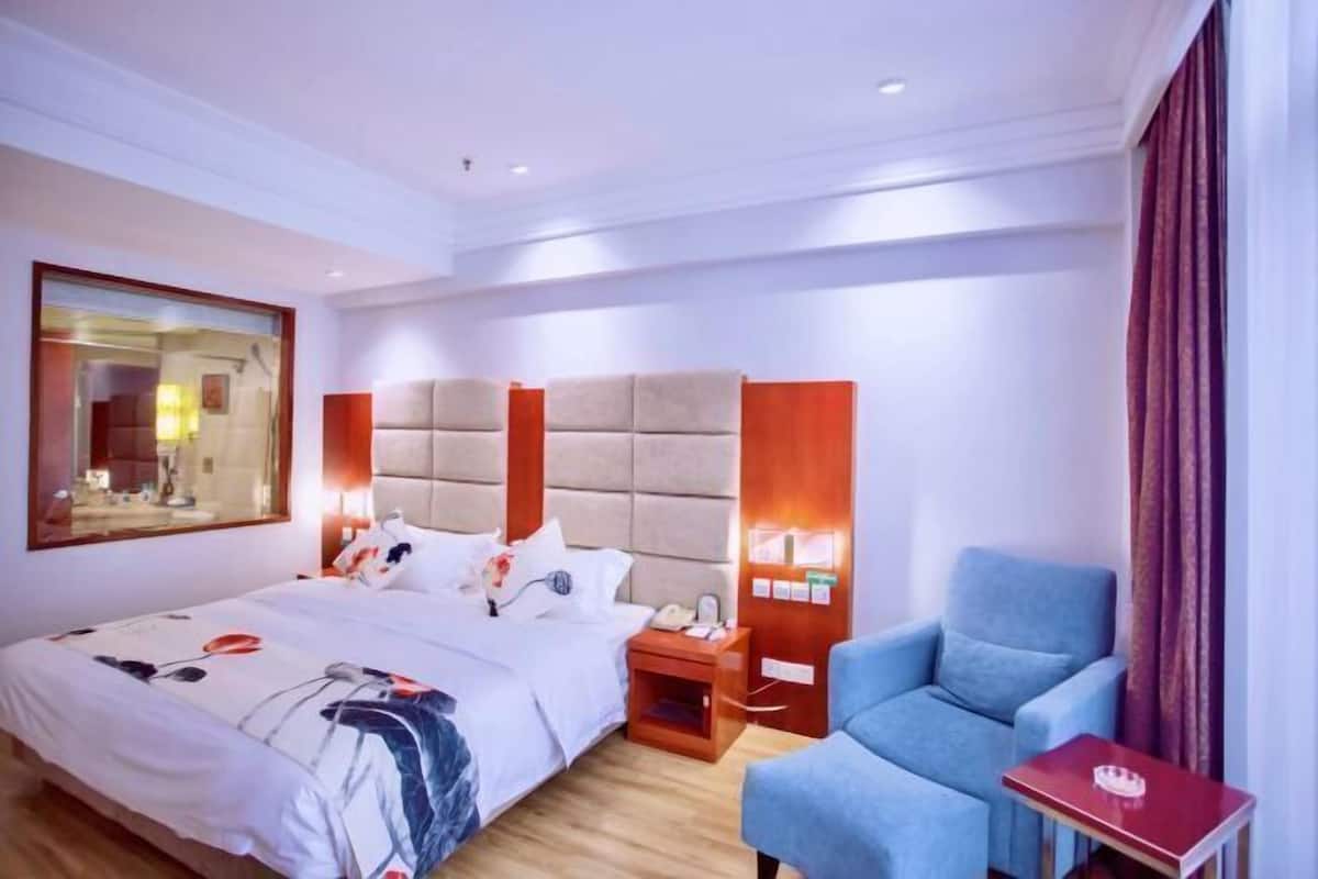 A hotel room with a large bed, a blue armchair, a red side table, and a mirror on the wall.