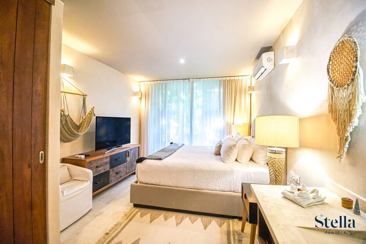 A hotel room with a bed, a TV, a desk, a chair, a lamp, and a wall-mounted air conditioner.