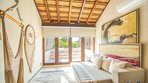 A cozy living room with a sofa, macramé wall hangings, a large abstract painting, and a view through glass doors to an outdoor seating area. - Eco-chic Design Condos Wrapped in Nature's Elements (Tulum)