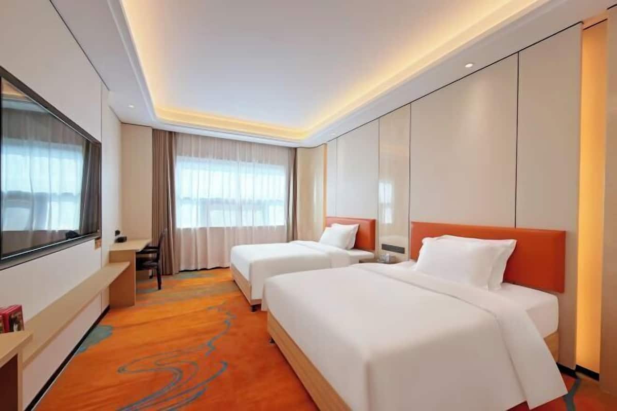 A hotel room with two beds, a TV, a desk, and a window with curtains.