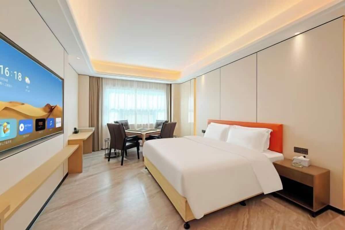 A hotel room with a bed, bedside table, nightstand, desk, chairs, and a TV displaying the time.