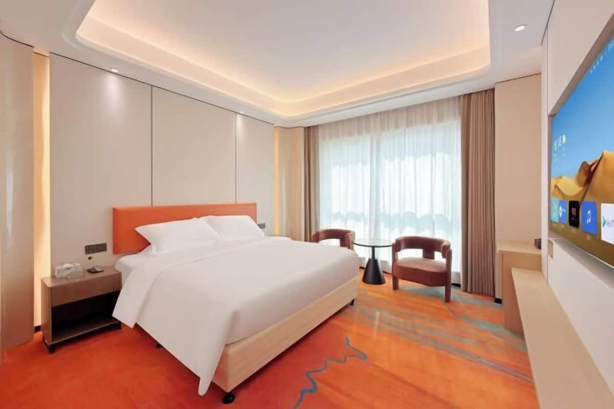 A hotel room with a bed, nightstand, chair, and a large window with curtains.