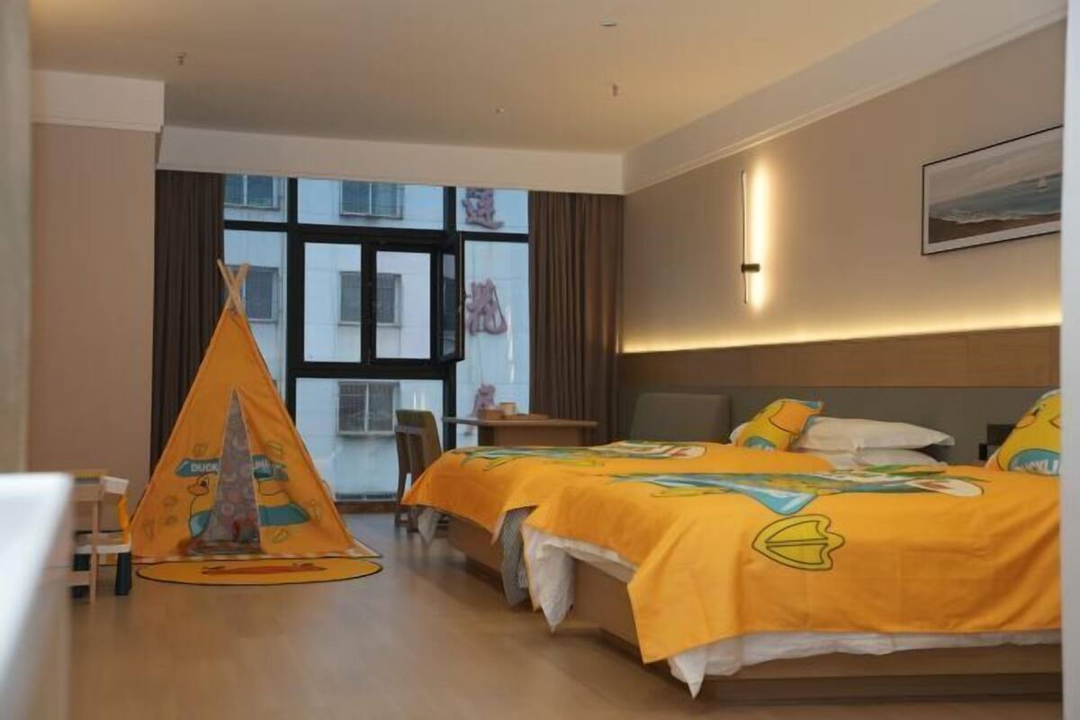A hotel room with two beds, a window with curtains, a small table, and a children's play tent.