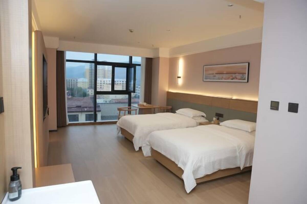 A hotel room with two beds, a desk, a chair, a window with a city view, and a painting on the wall.