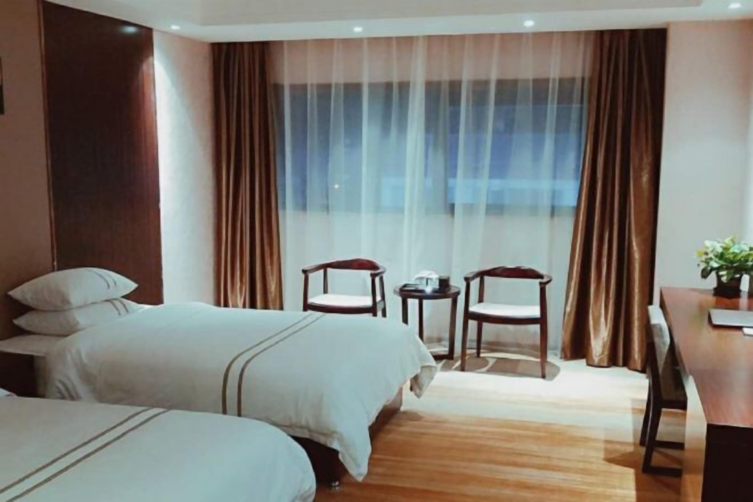 A hotel room with two beds, a dining table, chairs, a small table with a plant, and a large window with curtains.