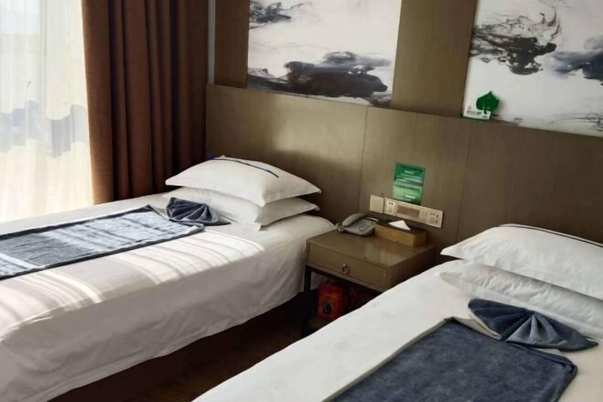 A hotel room with two beds, a nightstand with a phone and a green sign, and a window with curtains.