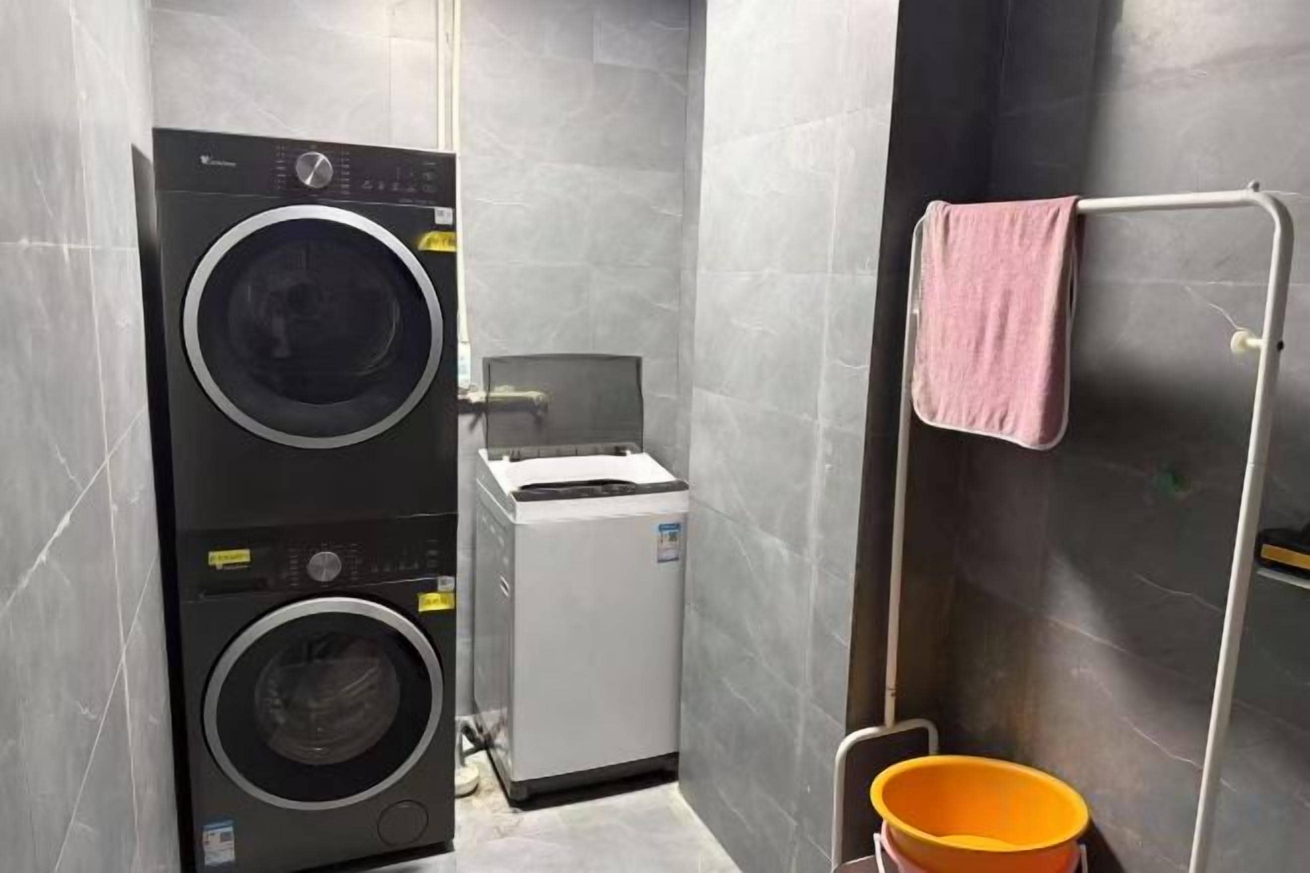 A compact laundry room with a stacked washer and dryer, a small white machine, and a metal drying rack with a pink towel.