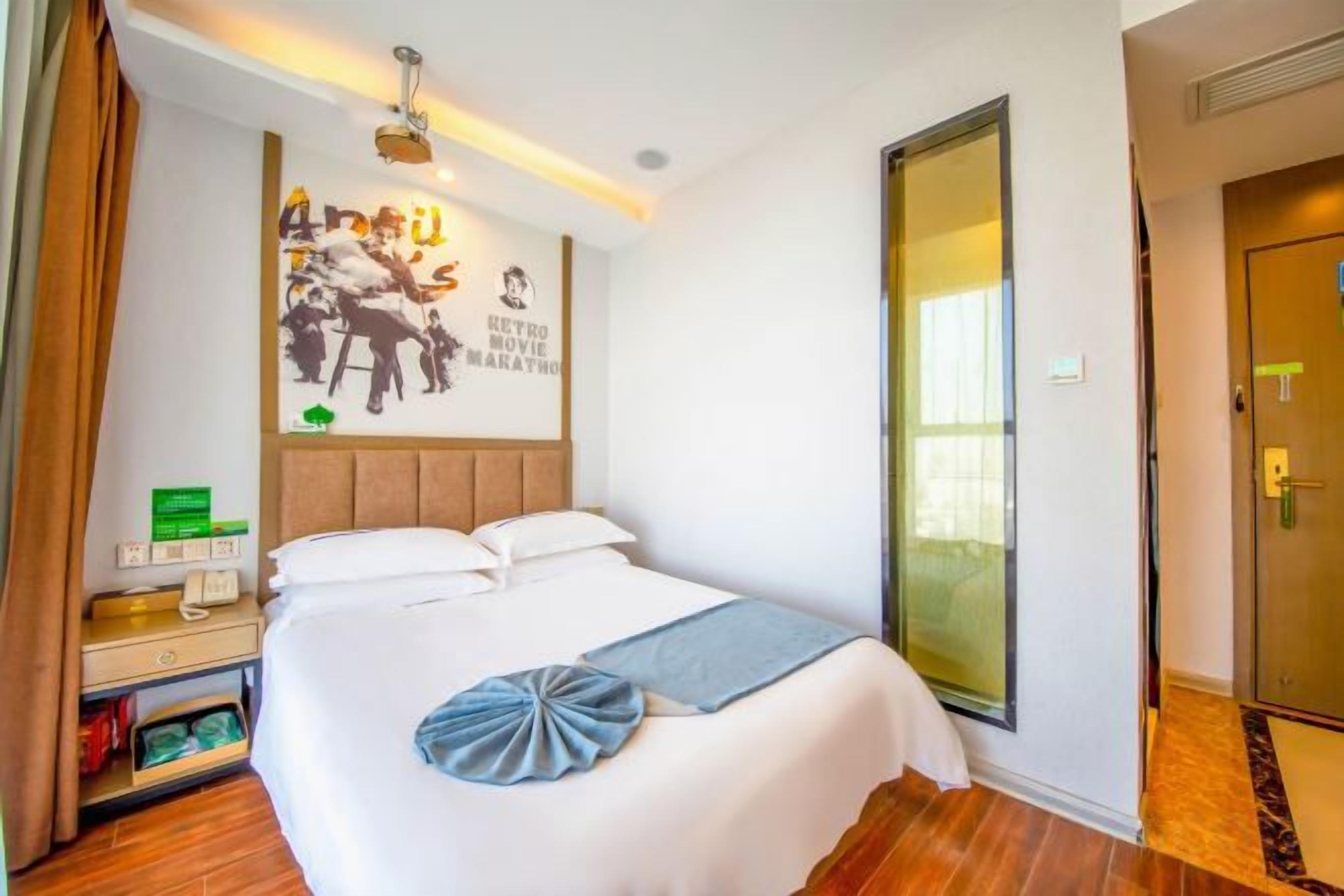 Habitación de hotel con una cama grande, una mesita de noche con una lámpara, un cartel en la pared y una puerta a la derecha.