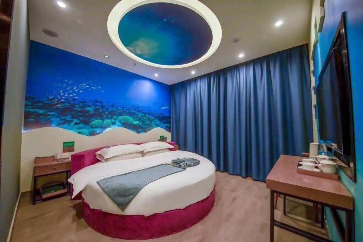 A room with a bed, a round ceiling light, a wall mural, a TV, and a small table with items.
