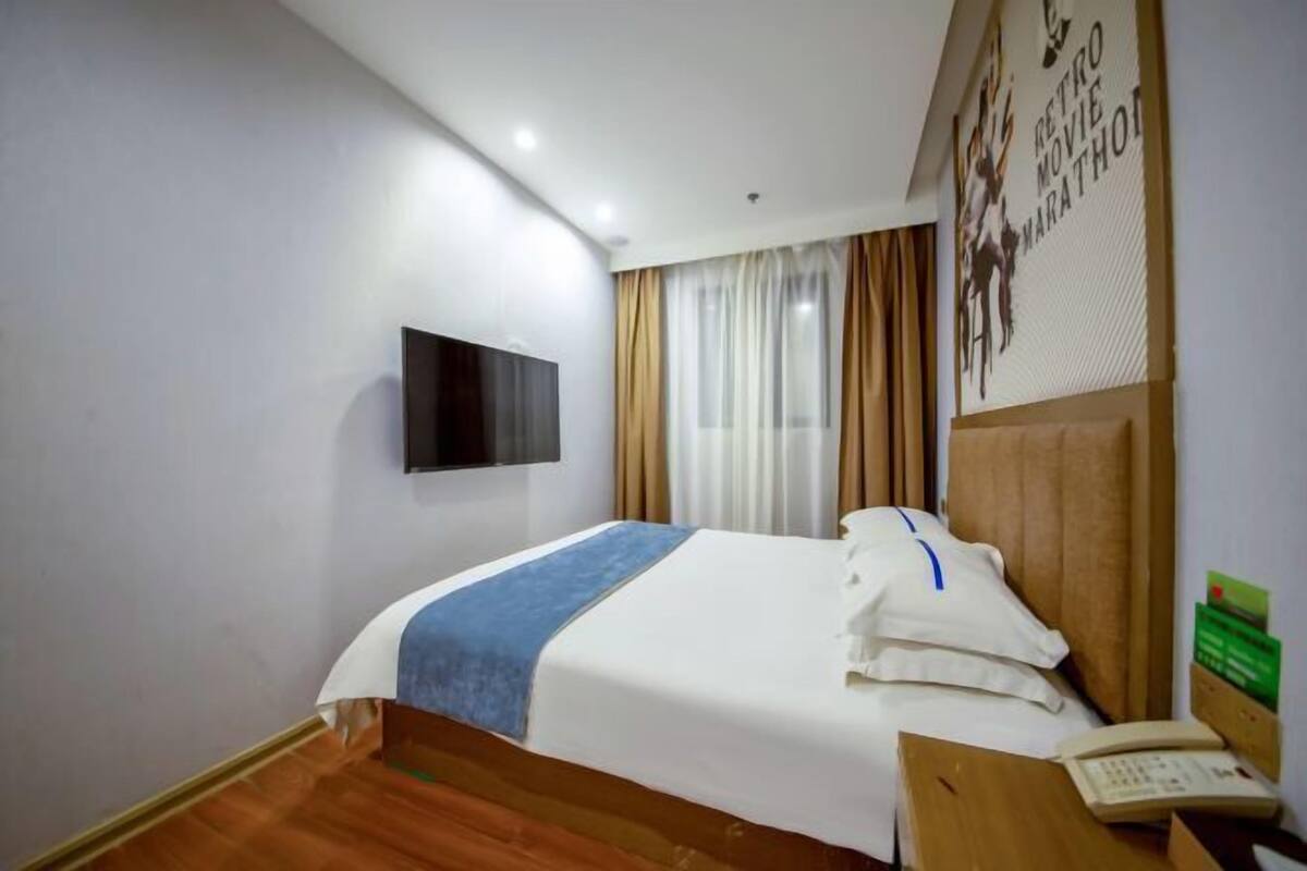 A hotel room with a bed, a flat-screen TV mounted on the wall, a wooden nightstand with a telephone, and a window with curtains.