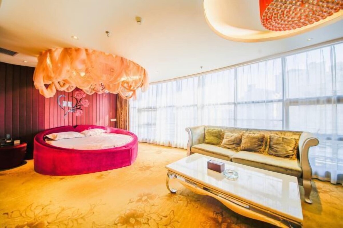 Un salon moderne avec un canapé circulaire rouge, un canapé beige, une table basse en verre et une grande fenêtre ornée de rideaux légers.
