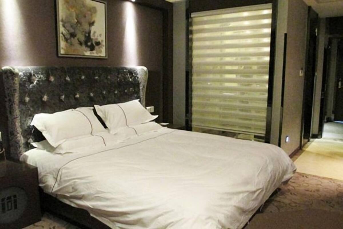 A hotel room with a large bed, a headboard, a window with blinds, and a painting on the wall.