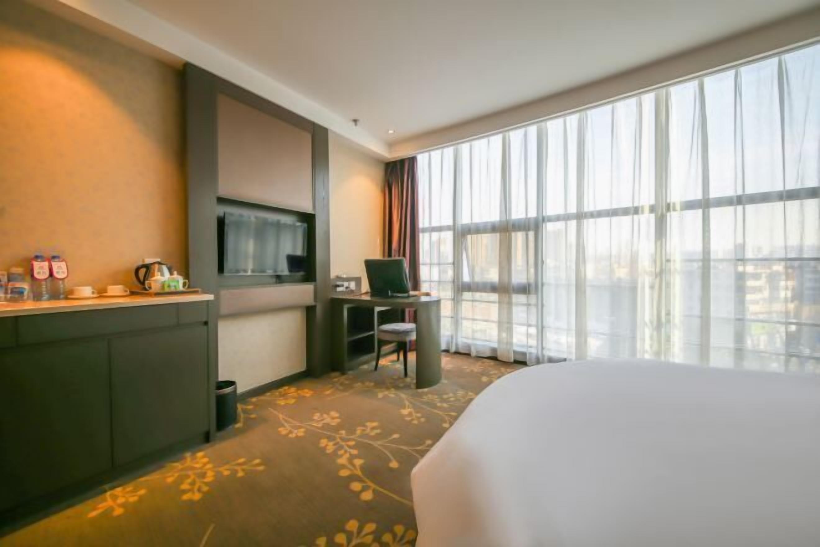 Habitación de hotel con una cama, un escritorio con una computadora portátil, un televisor y un ventanal con cortinas.