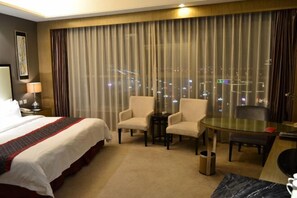 Habitación de hotel con una cama grande, dos sillones, una mesita y vistas de la ciudad de noche.