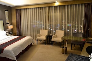Habitación de hotel con una cama grande, dos sillones, una mesita y vistas de la ciudad de noche.